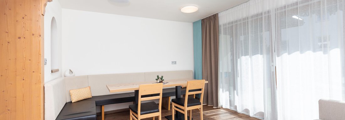 A bright and modern living room with wooden flooring and windows that provide plenty of daylight. There is a seating area with a table and chairs, as well as a cozy sofa. - Garni Stefani - Vent