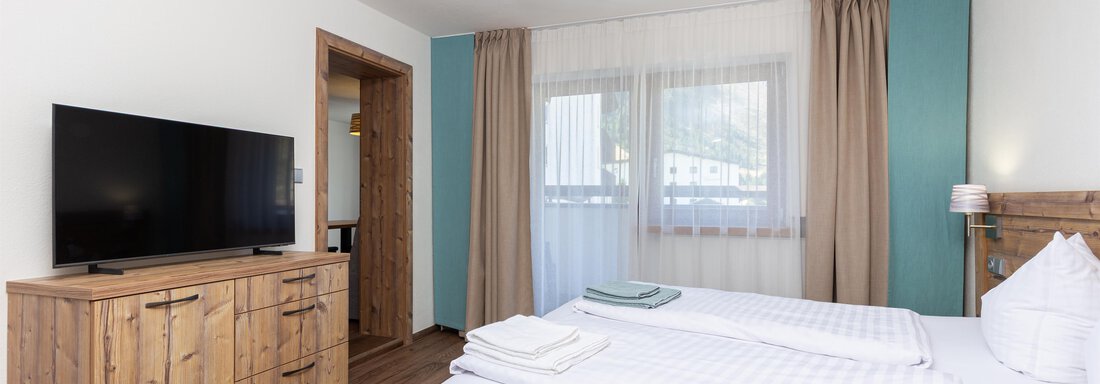 A modern bedroom with two single beds and a television. The furniture is made of wood and the room has large windows with curtains. - Garni Stefani - Vent