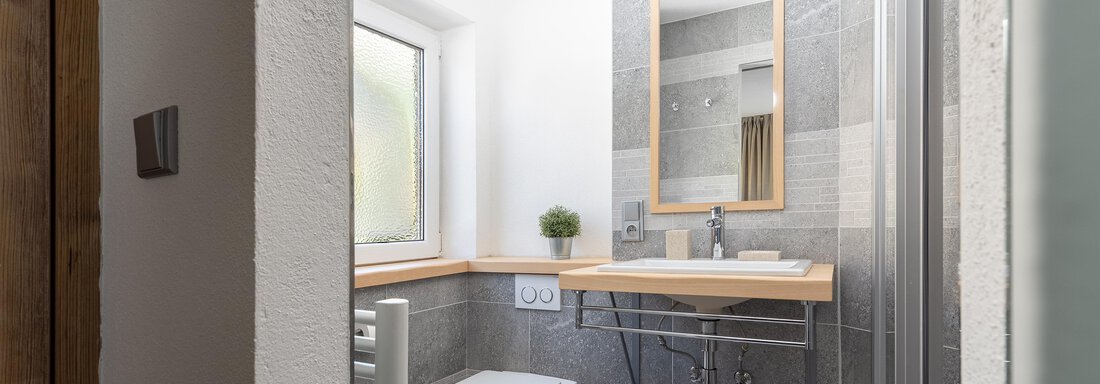A modern bathroom with gray tiles and a minimalist sink. There is a large mirror and plenty of natural light through the window. - Garni Stefani - Vent
