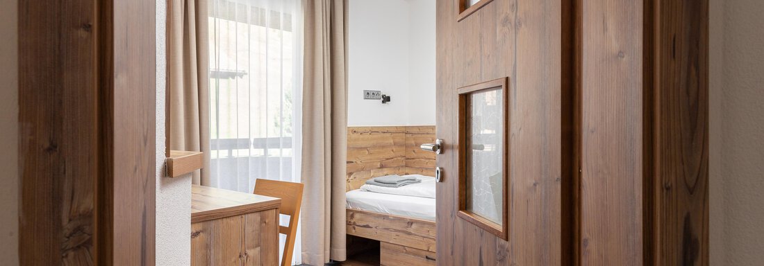 A cozy room with a wooden desk and a bed. The window lets in plenty of daylight and the walls are painted light. - Garni Stefani - Vent