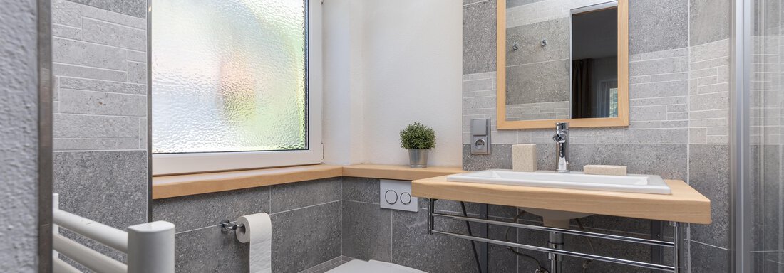A modern bathroom with gray tiles and a large mirror. It features a toilet, a sink, and natural light from a window. - Garni Stefani - Vent
