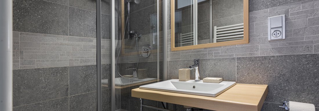 A modern bathroom with smooth gray tiles and a clear shower area. A sink with a wooden shelf and mirror creates an inviting atmosphere. - Garni Stefani - Vent