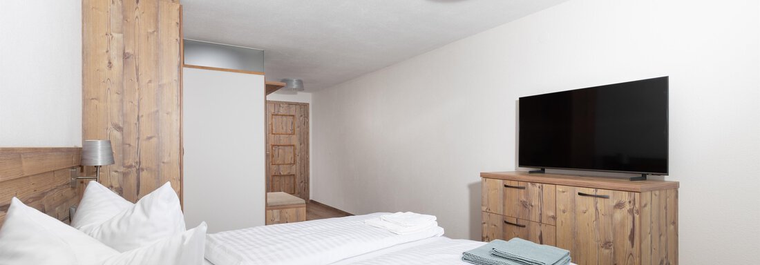 A modern bedroom with a large bed and two pillows. A television hangs on the wall and some wooden furniture gives the room a cozy atmosphere. - Garni Stefani - Vent