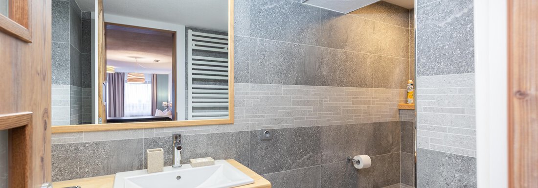 A modern bathroom with gray tiles and a wooden washbasin. The mirror reflects part of the room and there is a window for daylight. - Garni Stefani - Vent