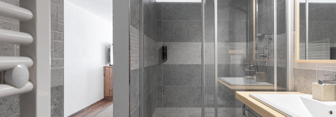A modern bathroom with a glass shower and a bright wood countertop. The walls are adorned with gray tiles and a large mirror light. - Garni Stefani - Vent