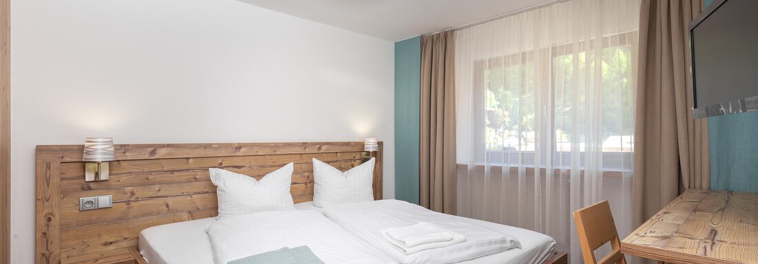 A modern bedroom with a wooden double bed and white bedding. The room has large windows with curtains and a small desk. - Garni Stefani - Vent