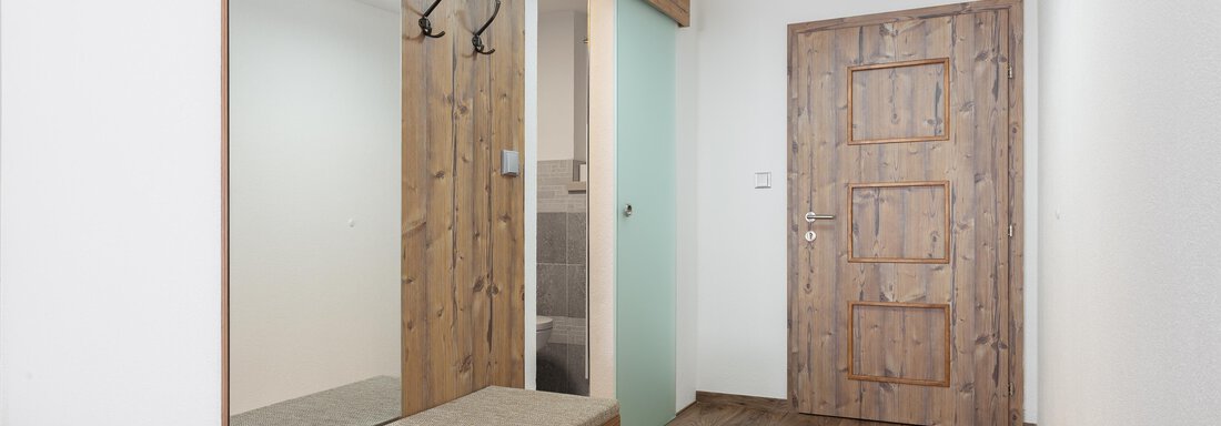 A modern entrance area with a wooden bench and wardrobe. The walls are light, and a glass door leads to another room. - Garni Stefani - Vent