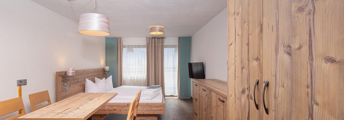 A cozy room with wooden furniture, a dining table, and a double bed. Large windows provide plenty of daylight. - Garni Stefani - Vent