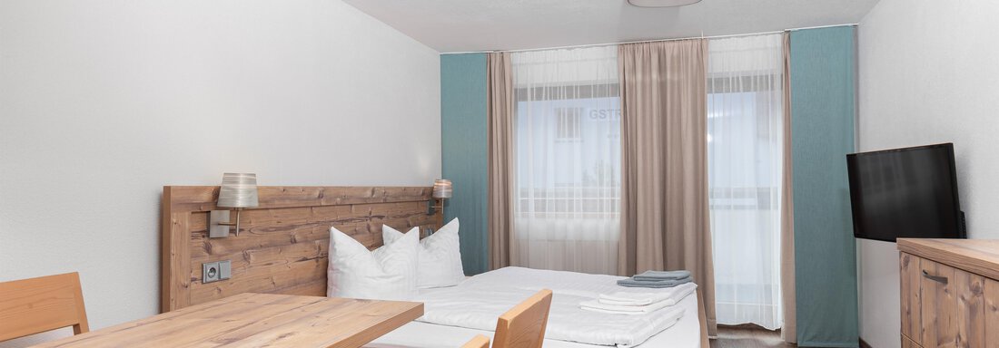 A modern room with a double bed and a wooden table. The room is bright and friendly, with a television and large windows. - Garni Stefani - Vent