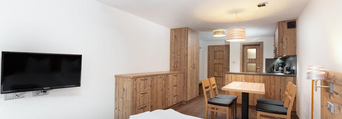 A modern room with wooden interiors and a double bed. There is a dining area with a table and chairs as well as a television on the wall. - Garni Stefani - Vent