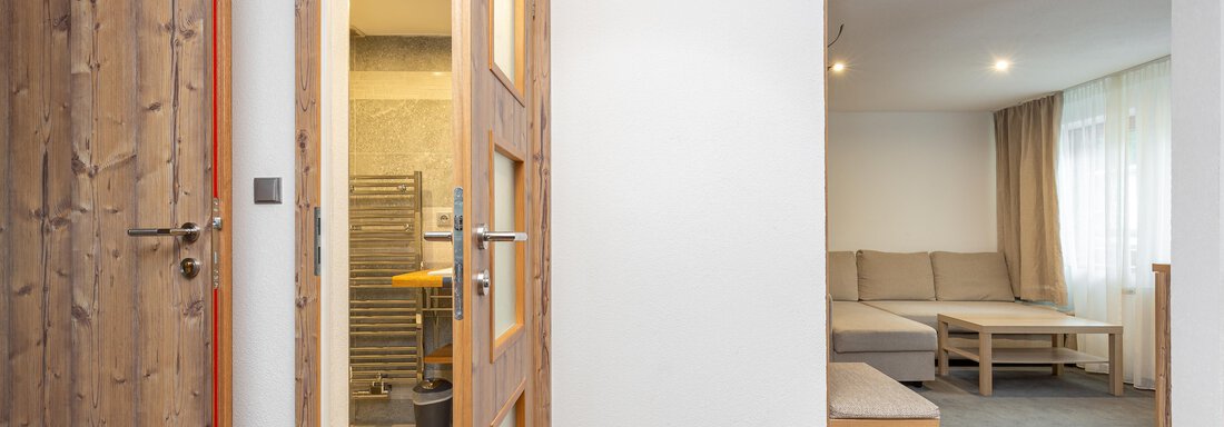 A bright room with wooden details and two open doors. On the right side, there is a cozy living area with a sofa and table. - Garni Stefani - Vent