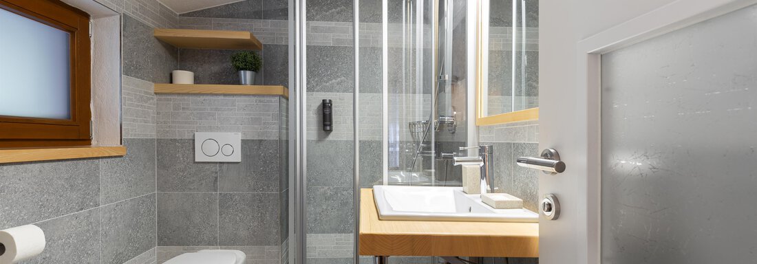 A modern bathroom with a shower and minimalist design. The walls are clad with gray tiles and there are wooden shelves. - Garni Stefani - Vent