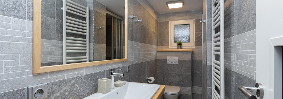 A modern bathroom with gray tiles and a large mirror. It features a sink, a toilet, and a towel radiator. - Garni Stefani - Vent