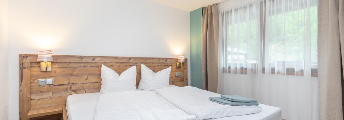 A cozy bedroom with a wooden double bed and white bed linens. Large windows let in plenty of light and offer a view of the outside world. - Garni Stefani - Vent