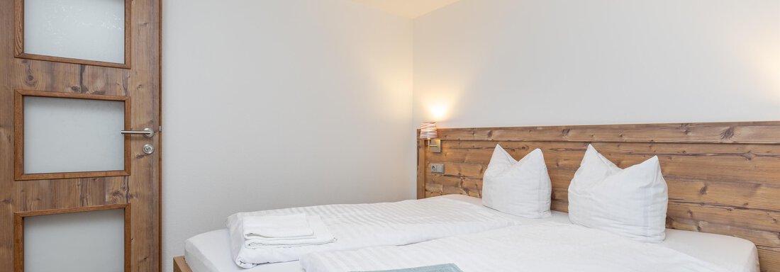 An inviting bedroom with a large bed and two pillows. The walls are light, and a wooden door leads outside. - Garni Stefani - Vent