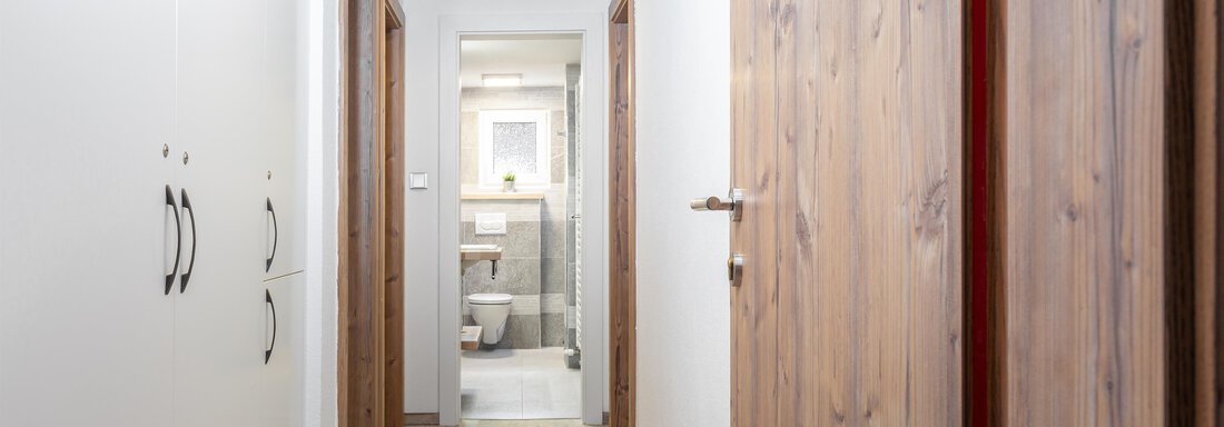 A bright hallway with wooden and white walls. At the end of the hallway, a bathroom can be seen. - Garni Stefani - Vent