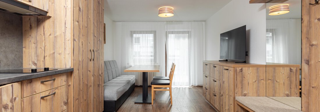 A modern apartment with wooden paneling and a cozy dining area. It features a sofa, a table, and a television in a bright room. - Garni Stefani - Vent