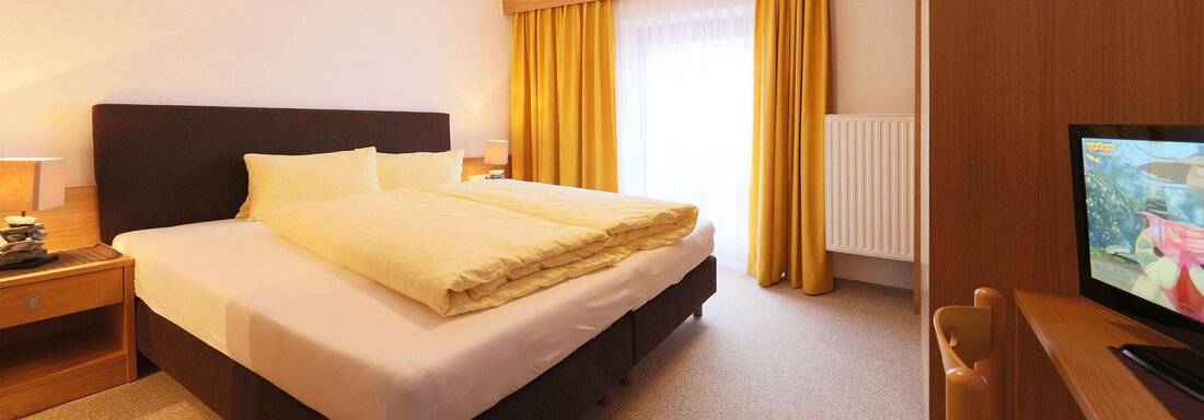 Zimmer_01 A cozy bedroom with a large bed and yellow curtains. There is a television and a bright, inviting space. - Gästehaus Edelweiss - Vent