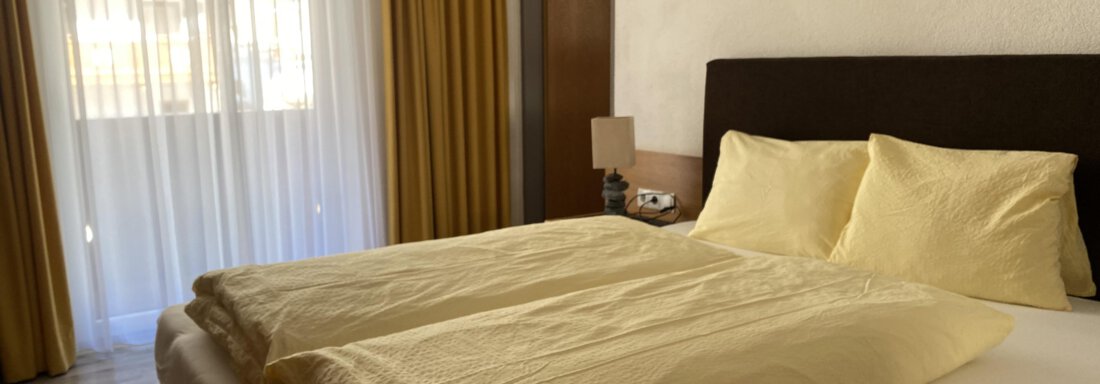 A bright bedroom with a large bed and yellow bedding. The windows are fitted with light curtains that let in daylight. - Gästehaus Edelweiss - Vent