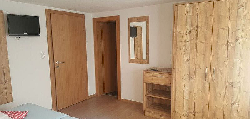 A simply furnished room with a wooden wardrobe and a small television. The walls are light and there is a large mirror. - Gästehaus Soldanella - Vent