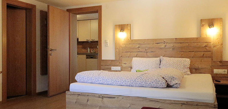 A cozy bedroom with a wooden bed and bright light. In the background, doors and a small kitchen are visible. - Gästehaus Soldanella - Vent
