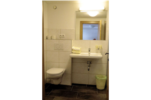 A modern bathroom with a white toilet, a hand sink, and a large mirror. The walls are light and tiled, and there are fresh towels on the sink. - Gästehaus Soldanella - Vent