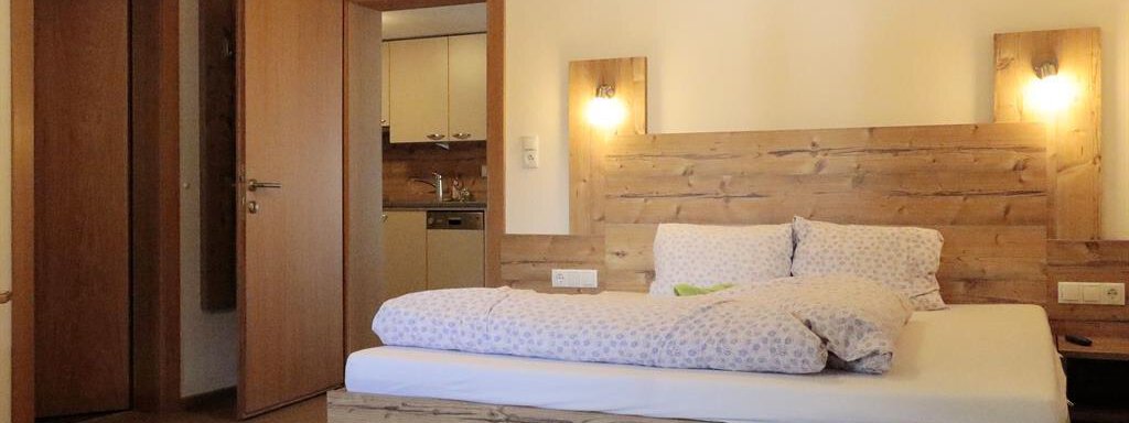 A cozy bedroom with a wooden bed and gentle lighting. In the background, a kitchen and two doors are visible. - Gästehaus Soldanella - Vent
