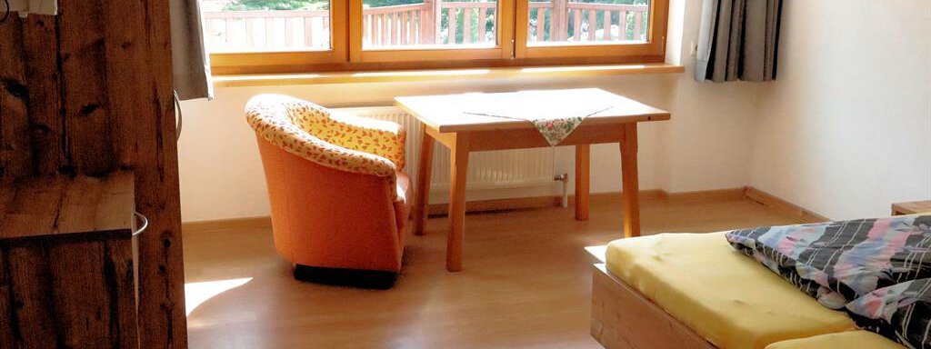 A simple room with wooden furniture, a bed, and a table. Large windows offer a view of nature outside. - Gästehaus Soldanella - Vent