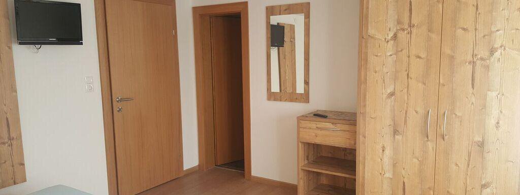 A cozy bedroom with wooden furniture and a small television on the wall. There is a door to another room and a mirror next to a nightstand. - Gästehaus Soldanella - Vent