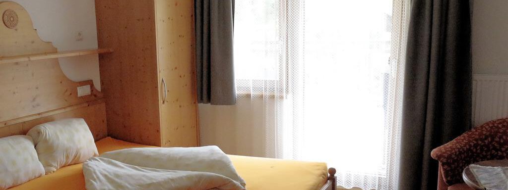 A cozy bedroom with a large bed and wooden furniture. Gray curtains hang in front of the window, and there is a comfortable seating area. - Gästehaus Soldanella - Vent