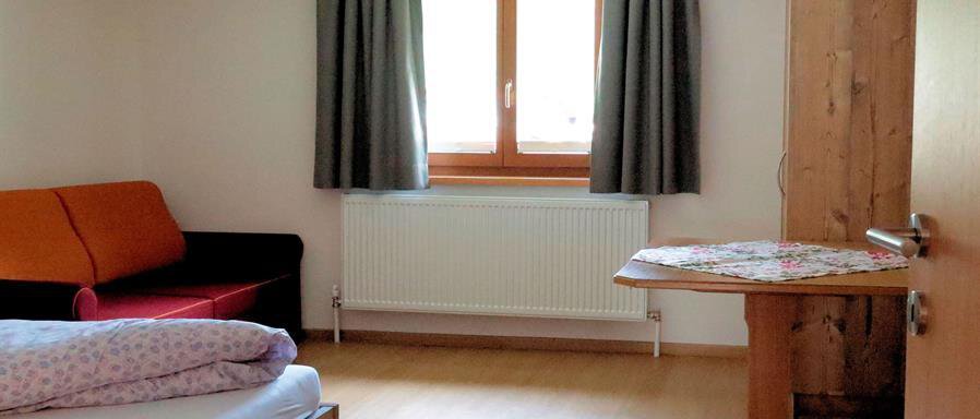 A simple room with a bed, a table, and a couch. The room has a window with curtains and a wooden wardrobe. - Gästehaus Soldanella - Vent