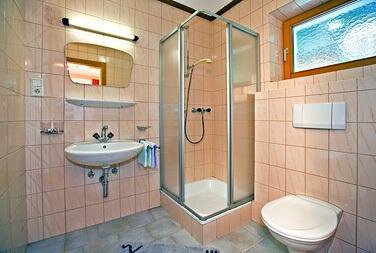 A small bathroom with pink tiles, shower, sink, and toilet. There is a window that lets in daylight. - Haus Weisskogelblick - Vent