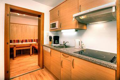 A modern kitchen with wooden cabinets and a stove. In the background, there is a dining area with a wooden bench. - Haus Weisskogelblick - Vent