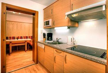 A modern kitchen made of wood with a stove, microwave, and a sink. In the background, there is a dining area with a table and benches. - Haus Weisskogelblick - Vent