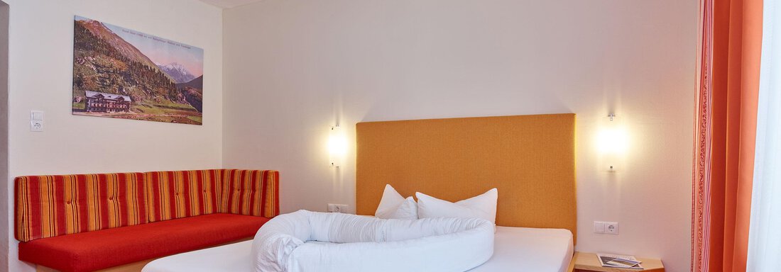 A cozy bedroom with a double bed and a red bench. The walls are light, and there is a picture of a mountain in the background. - Hotel Vent - Vent