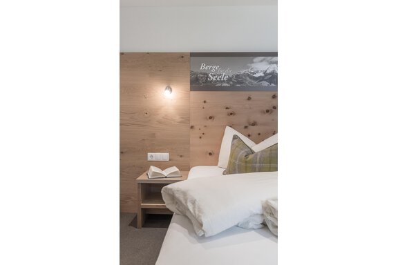 A cozy bedroom with a wooden wardrobe and a modern bedside table. On the bed are pillows in soft colors and an open book. - Garni Ötztal ROOMS & APARTS - Zwieselstein