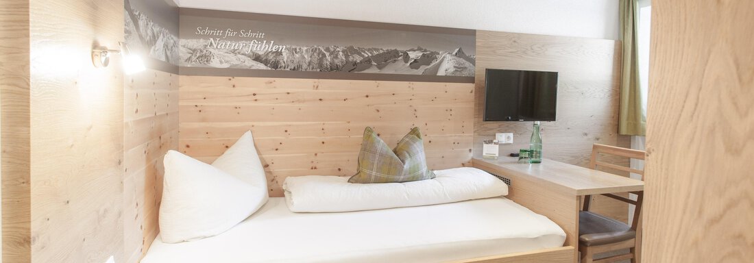 Einzelzimmer | © Contrast Group, Michaela Jörg (CC C) A modern room with wooden paneling and clean lines. It contains a single bed, a television, and a small desk. - Garni Ötztal ROOMS & APARTS - Zwieselstein