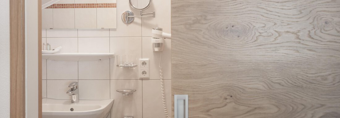 A modern bathroom with white tiles and a washbasin. The wooden door gives the room a warm atmosphere. - Garni Ötztal ROOMS & APARTS - Zwieselstein