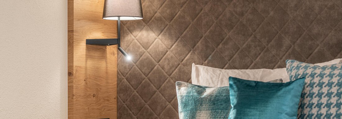 A modern bedroom with a comfortable bed and decorative pillows. The walls are adorned with a stylish, upholstered design and illuminated by a pleasant lamp. - Garni Ötztal ROOMS & APARTS - Zwieselstein