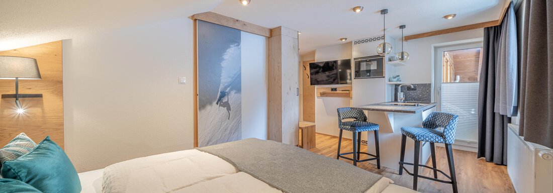 A modern room with two beds and minimalist decor. In the background, there is a kitchenette with bar stools. - Garni Ötztal ROOMS & APARTS - Zwieselstein