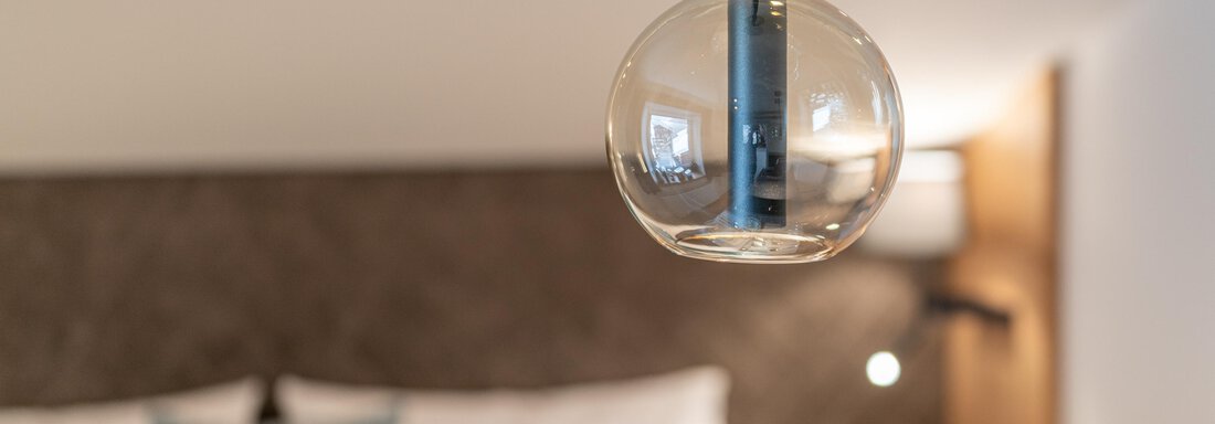 A modern ceiling lamp hangs over a cozy bedroom. The bedding is decorated with blue and white pillows. - Garni Ötztal ROOMS & APARTS - Zwieselstein