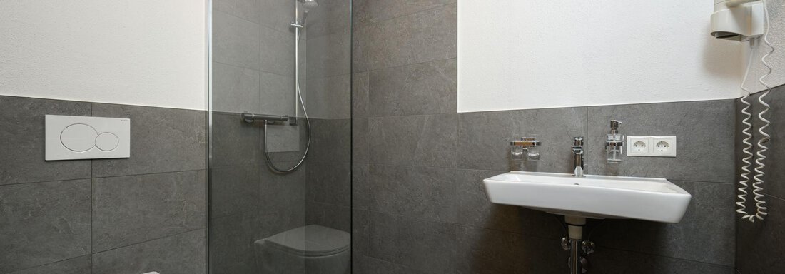 A modern bathroom with a glass shower, a sink, and a toilet. The walls are clad with gray tiles. - Gästehaus Judith - Zwieselstein