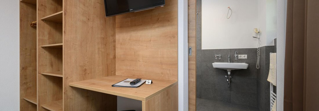 A modern room with a wooden shelf, a desk, and a television. In the background, a bathroom with a sink is visible. - Gästehaus Judith - Zwieselstein