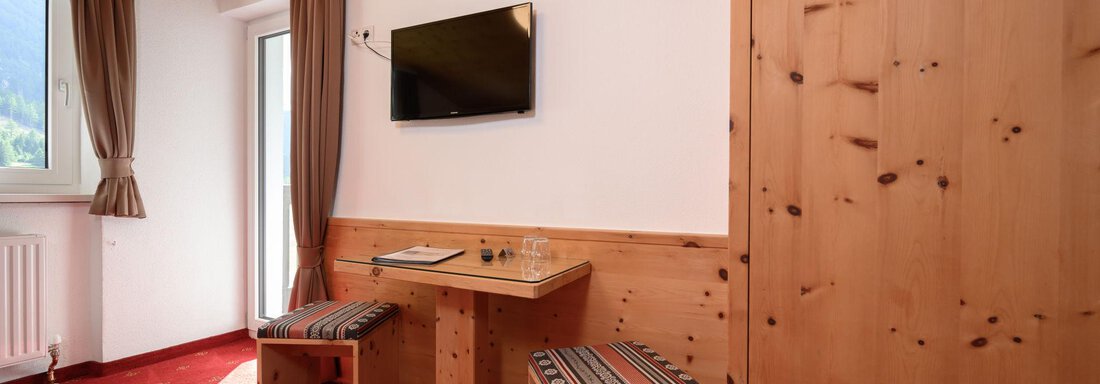 A cozy room with wooden decor, a television, and a small table. There are also two chairs and a window with a view outside. - Gästehaus Judith - Zwieselstein