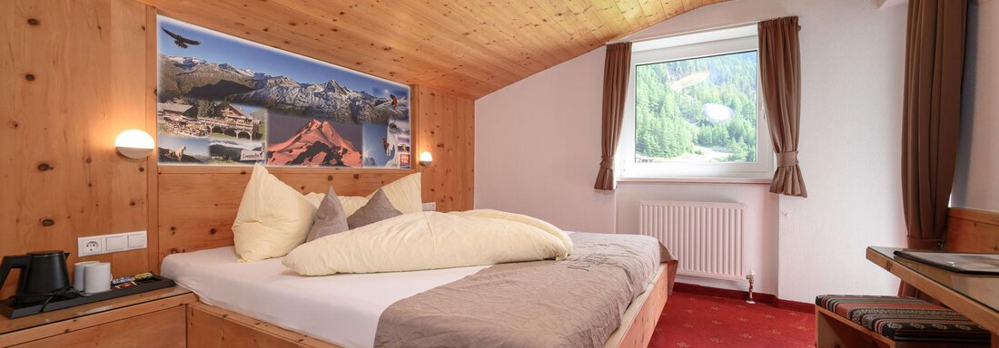 A cozy bedroom with wooden paneling and a large window. The bed is inviting, equipped with cushions and blankets. - Gästehaus Judith - Zwieselstein