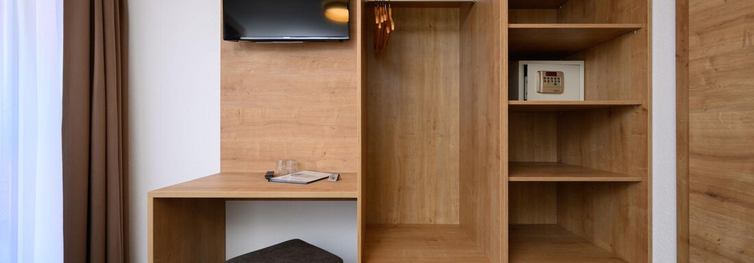 A modern room with a wooden closet and a desk. There is a television on the wall and a small stool stands next to it. - Gästehaus Judith - Zwieselstein