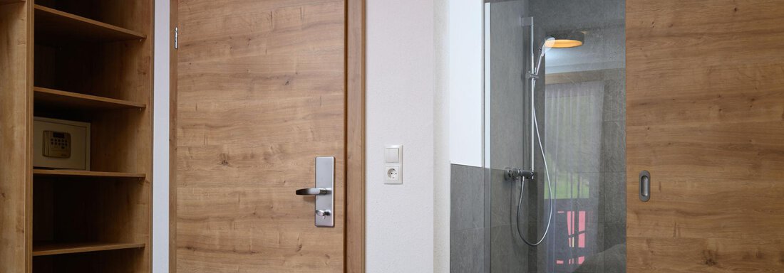 A modern room with a wooden shelf and a wooden door. In the background, a bathroom with a shower can be seen. - Gästehaus Judith - Zwieselstein
