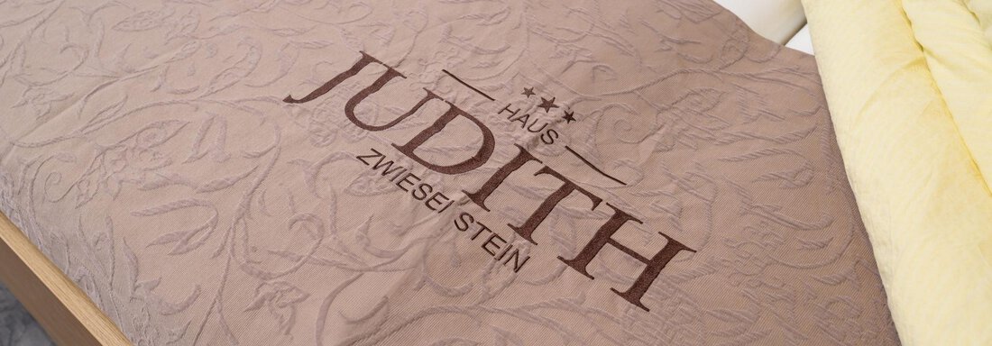 A bed with elegant bedding and a slightly textured surface. The "Judith" logo is prominently displayed in the center of the bed sheet. - Gästehaus Judith - Zwieselstein