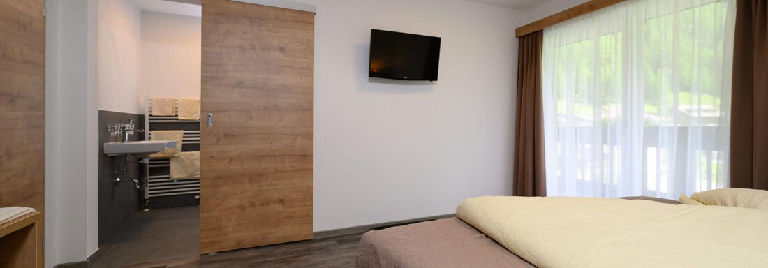 A modern bedroom with two beds and a flat-screen TV. In the background, an adjoining bathroom is visible. - Gästehaus Judith - Zwieselstein