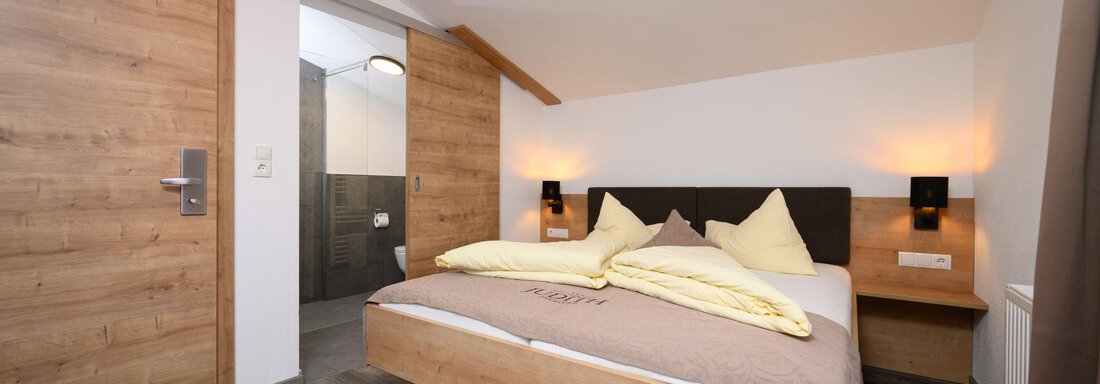 A modern bedroom with a double bed and wooden details. The walls are bright, and there is a window that lets in natural light. - Gästehaus Judith - Zwieselstein